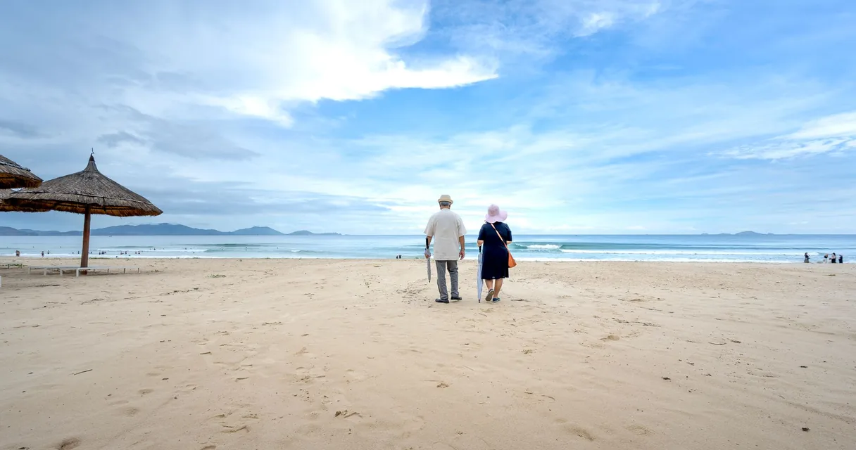 Casal idoso caminhando de mãos dadas na areia de uma praia sob céu azul